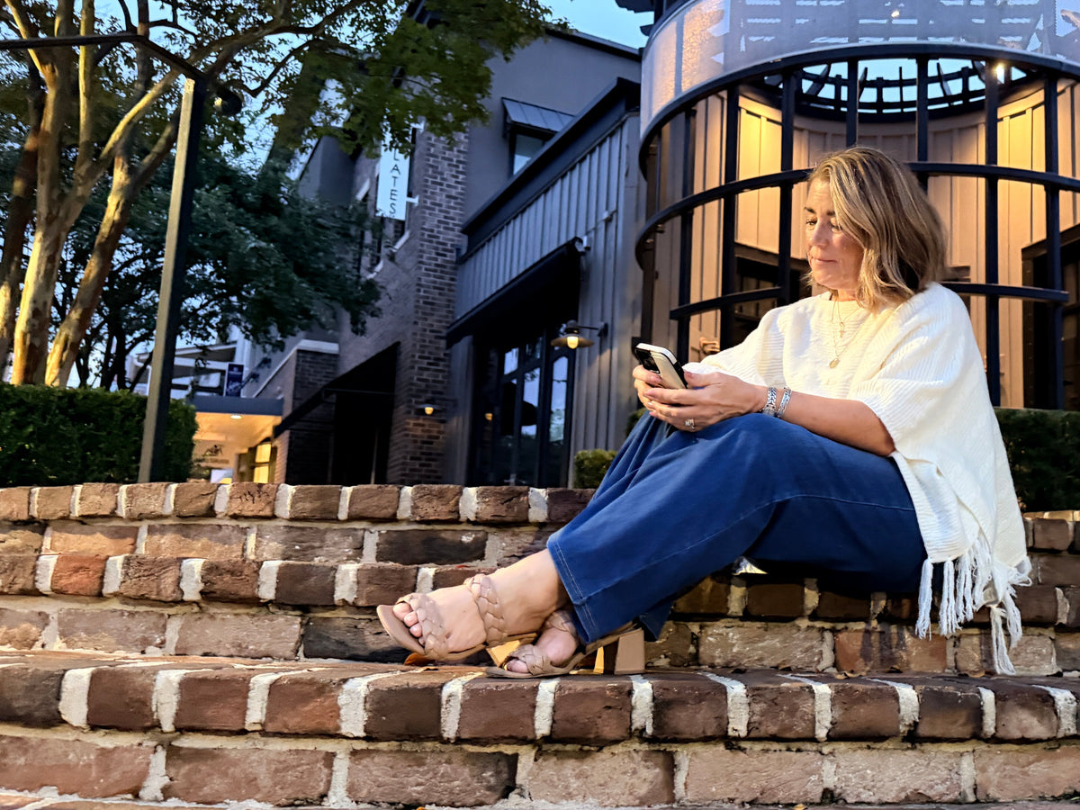 A woman sits on outdoor brick steps in front of a modern building, holding a phone. She is wearing a white fringed top, blue pants, and beige sandals, with a green tote bag resting on the steps nearby.